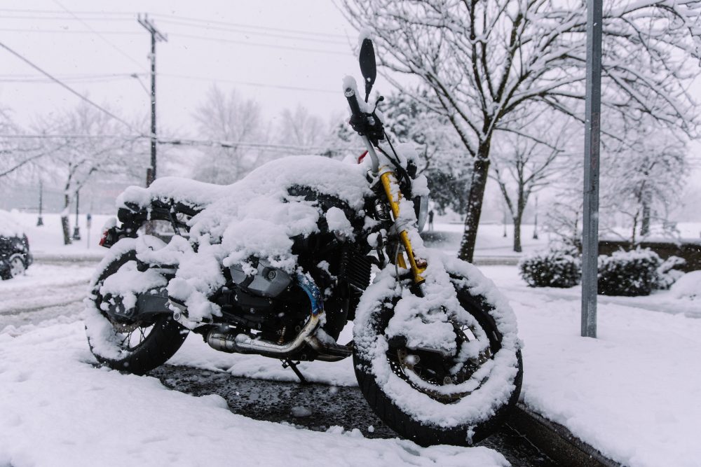 Motocykl cały w śniegu, pozostawiony na parkingu bez żadnego przykrycia, co podkreśla potrzebę odpowiedniego zimowania motocykla, aby uniknąć jego uszkodzeń.